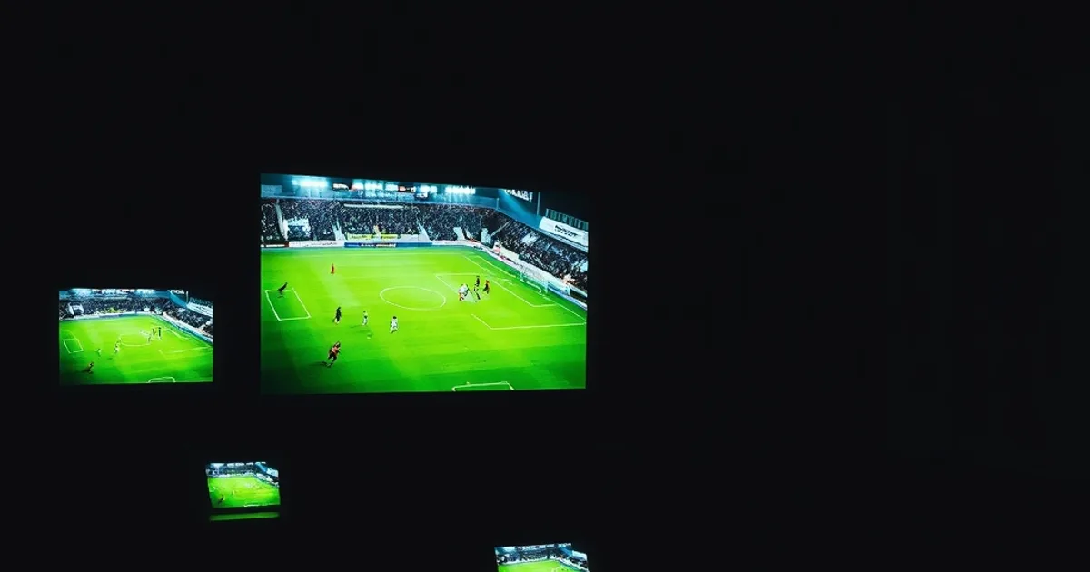 Salón oscuro por la noche iluminado por un televisor, una tablet y un móvil mostrando partidos de fútbol en directo al mismo tiempo, ambiente cinematográfico